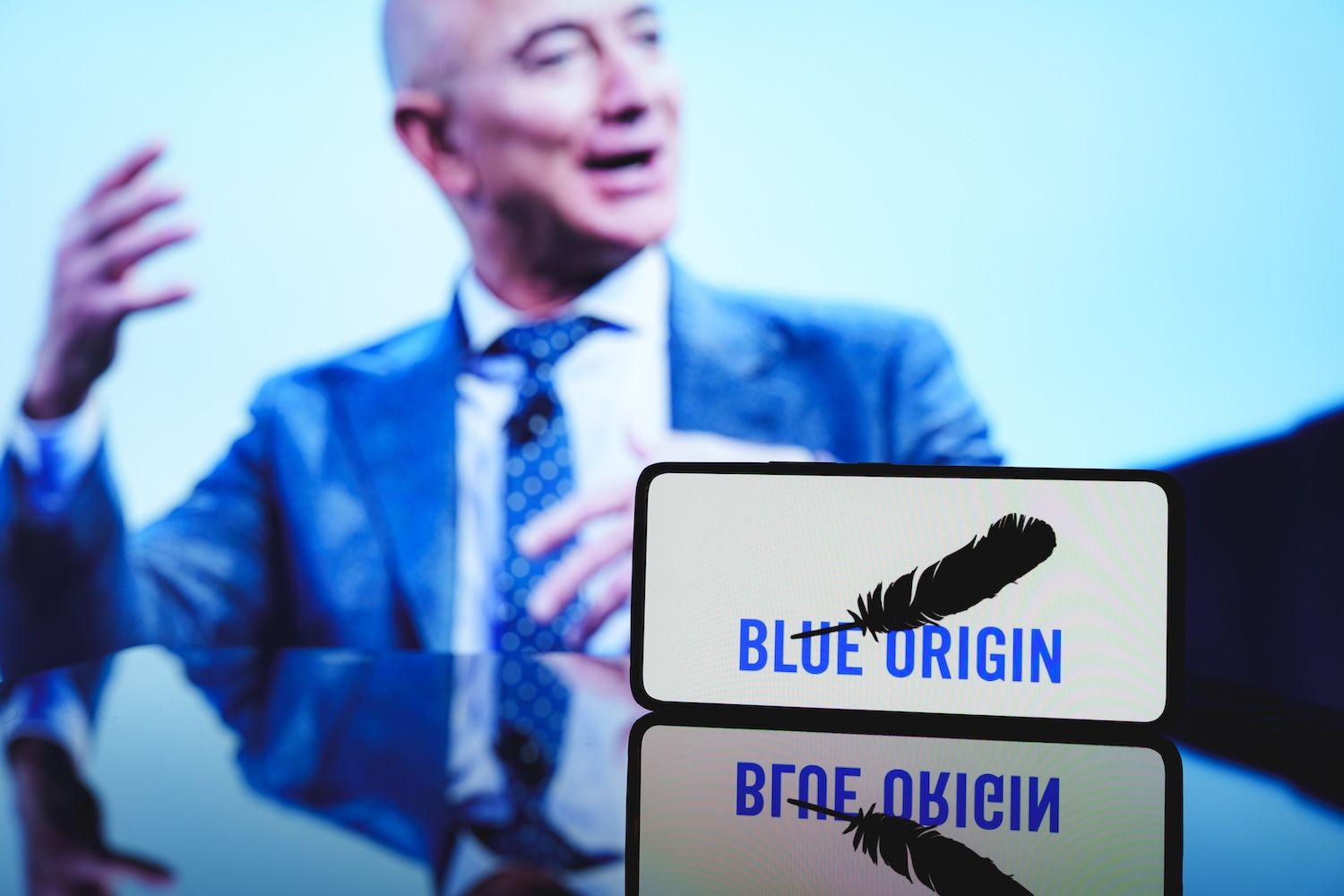 Jeff Bezos speaking with the Blue Origin logo displayed on a smartphone screen in the foreground.
