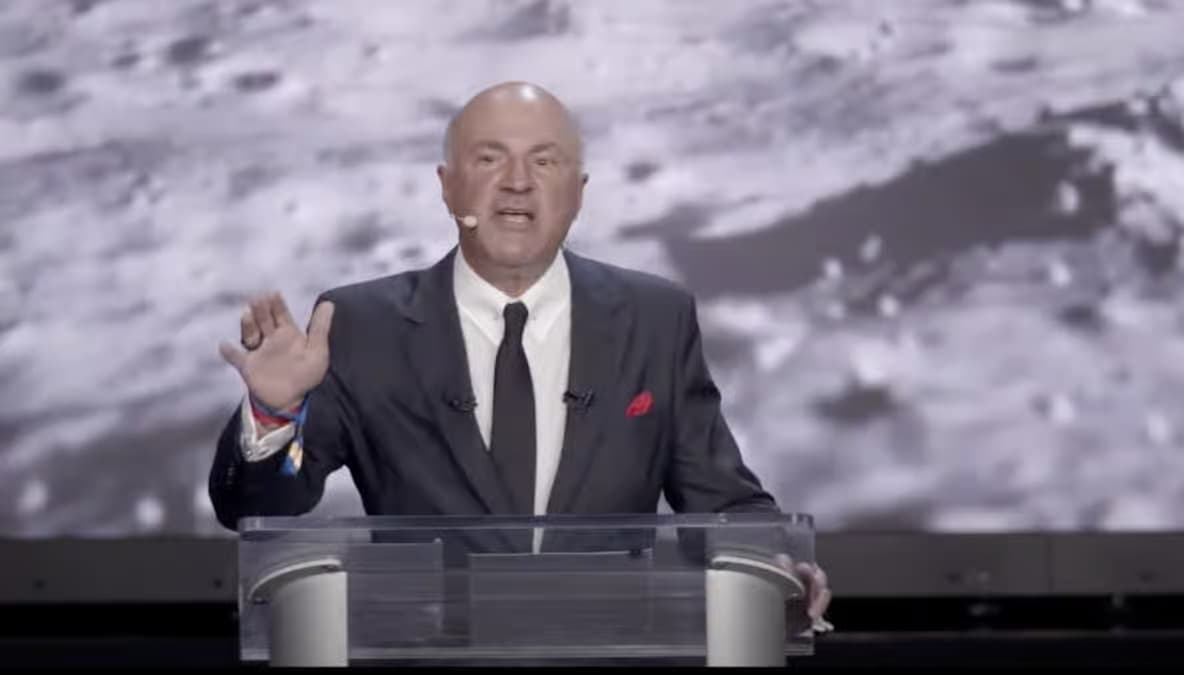 Man in a dark suit with red pocket square speaking at a podium with a lunar surface image projected behind him