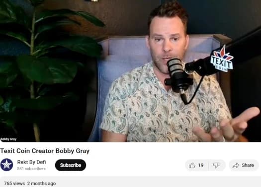 Screenshot of Bobby Gray, creator of Texit Coin, speaking into a microphone labeled TEXIT COIN during an interview with a plant in the background