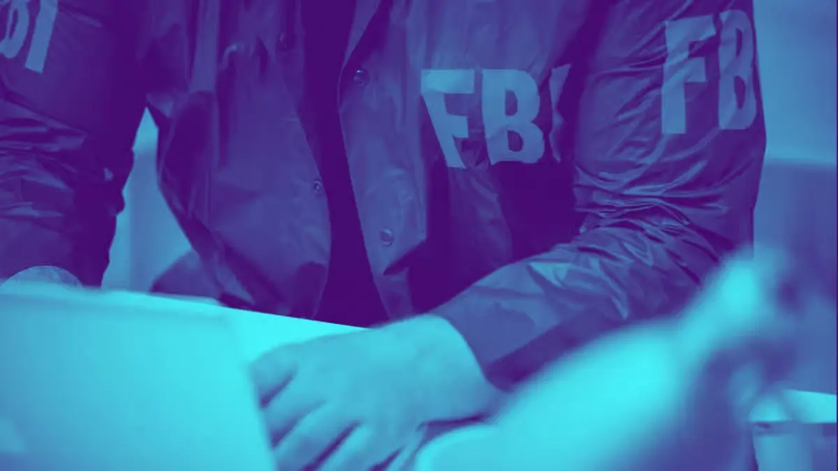 A person wearing a jacket with FBI printed on the sleeves while working on a laptop, stylized in a purple and blue duo-tone filter to represent cybercrime investigation and the 2025 crypto scam report.