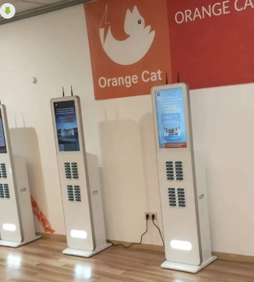 Three white Orange Cat power bank rental kiosks with digital screens placed against a wall with brand banners.