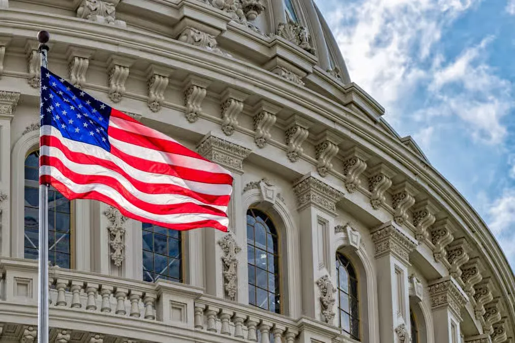 American flag waving in front of the US Capitol building under a blue sky in Washington, D.C.