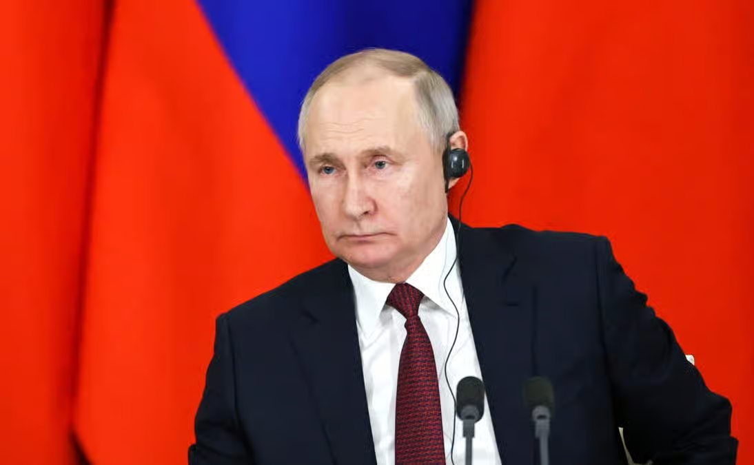 Portrait of Russian President Vladimir Putin wearing a dark suit, red tie, and an earpiece, sitting in front of the Russian flag.