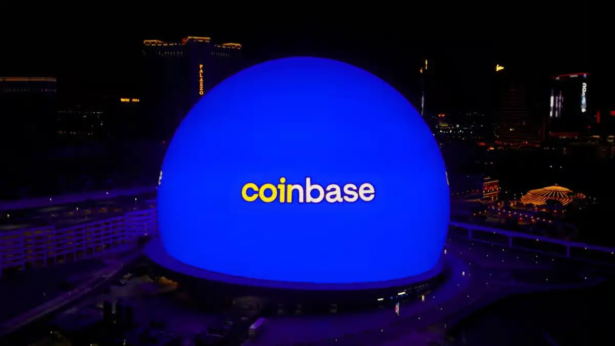 Large glowing blue Sphere building at night in Las Vegas displaying the Coinbase logo in yellow and white, with the Palazzo hotel and city lights visible in the background.