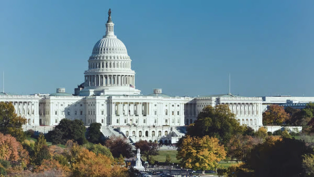 Exterior view of the US Capitol Building dome in Washington DC surrounded by autumn trees under a clear blue sky.