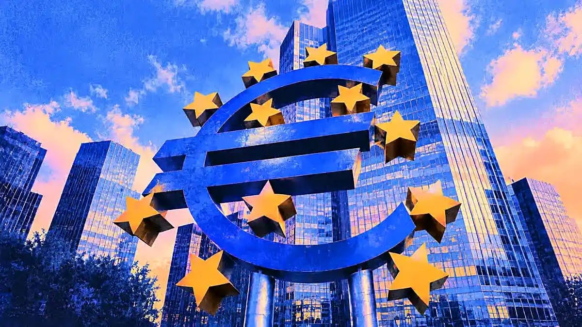Large blue Euro currency sculpture with yellow stars standing in front of modern financial skyscrapers under a vibrant sky.